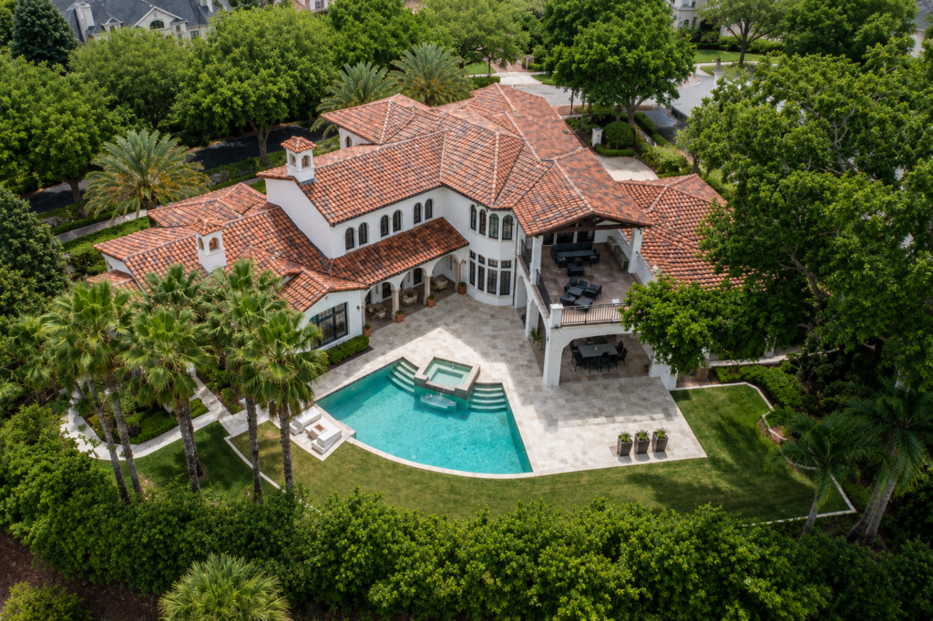 Aerial view of a Mediterranean estate with a clay barrel tile roof, courtyard pool, and paver deck framed by palms