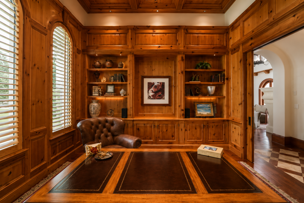Wood-paneled study with built-in bookshelves, a coffered ceiling, an arched window, and a tufted leather chair