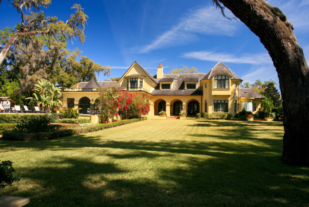 Rear elevation of a French estate home with steep hipped roofs, dormers, and a turret, set on a lawn beneath live oaks