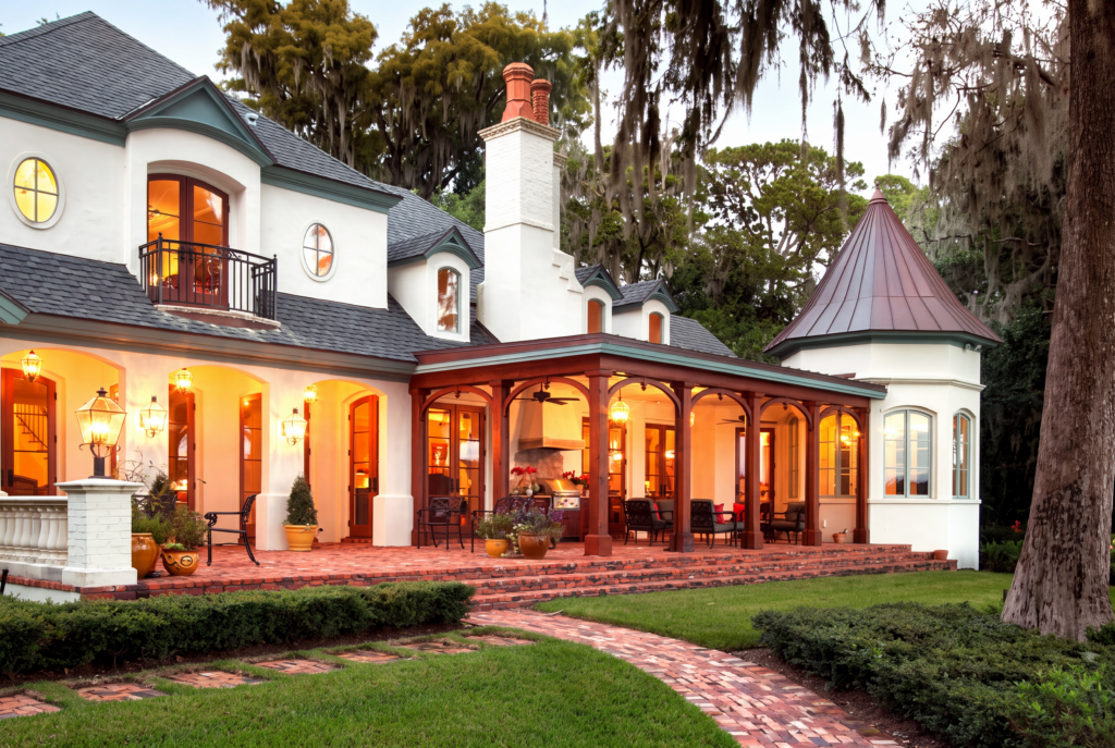 Rear elevation at dusk with a covered veranda, brick chimneys, a turret, and a brick paver walk beneath live oaks