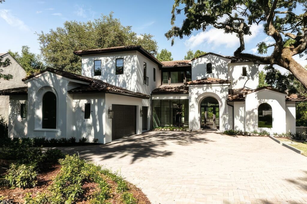 Front elevation with white stucco, clay tile roof, arched openings, and a paver motor court beneath a live oak