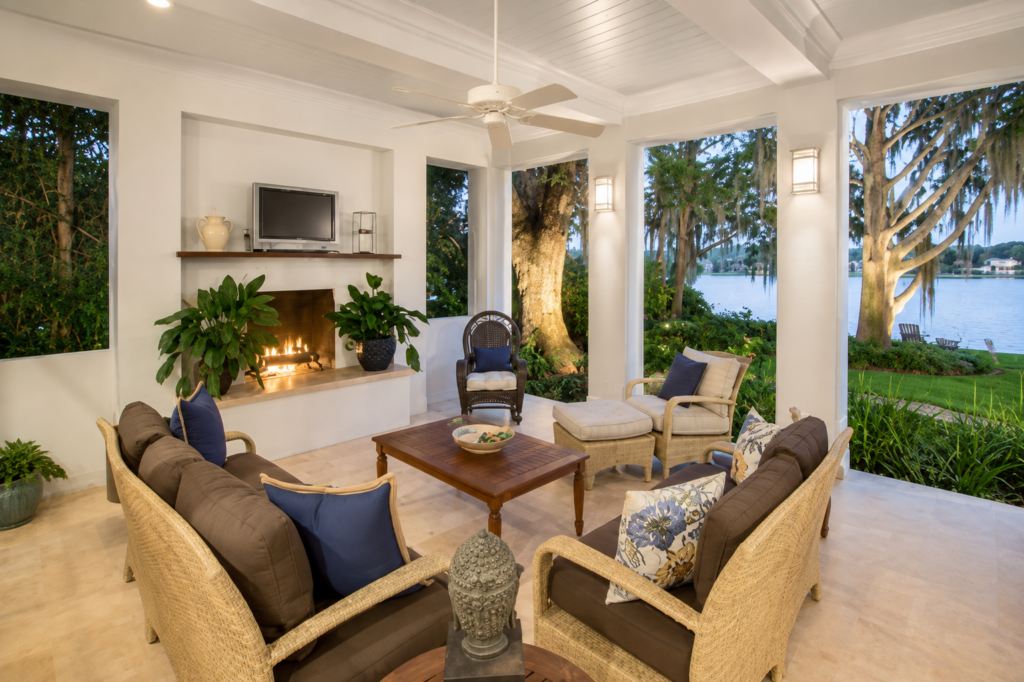 Covered rear porch with a fireplace, plank ceiling, and wicker seating looking out through live oaks to the lake