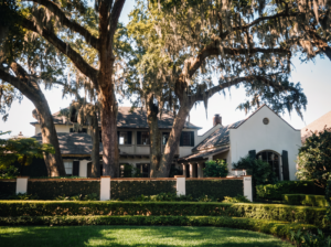 Front elevation behind a stucco garden wall and clipped hedge, set under live oaks draped in Spanish moss