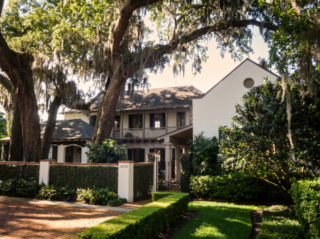 Street view of a Creole-influenced home with a stucco wall, iron gate, and second-story gallery beneath moss-draped oaks